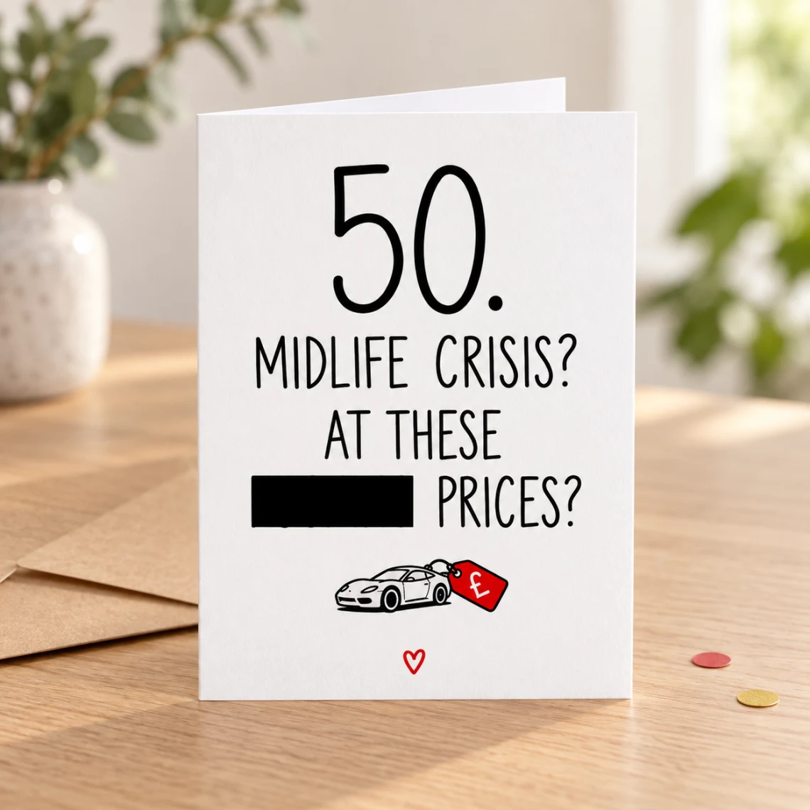 Lifestyle mock-up of a standing 50th birthday card about midlife-crisis prices, with one word covered by a black bar and a sports-car price-tag doodle. On a wooden table beside a kraft envelope and red/gold confetti.