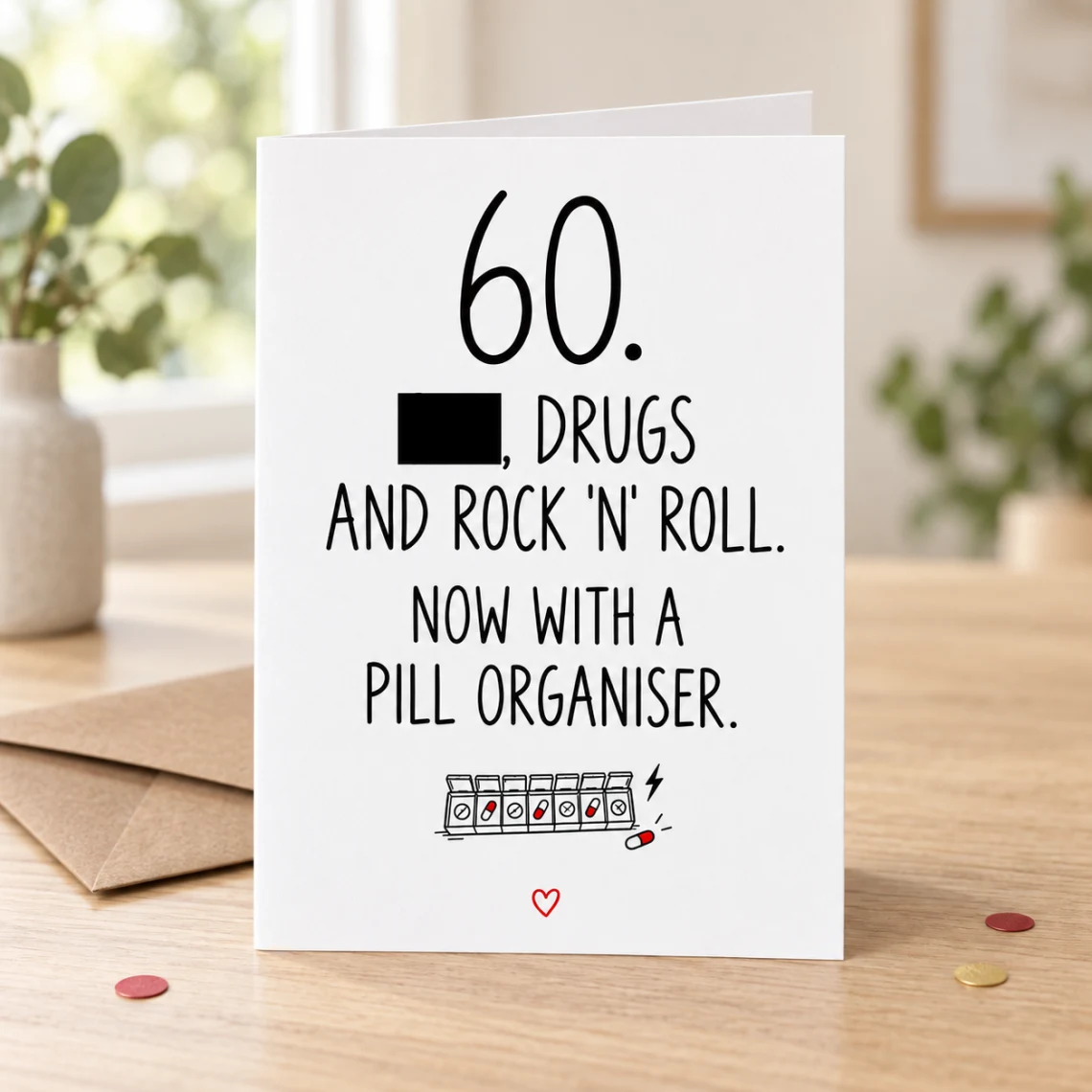 Lifestyle mock-up of a standing 60th birthday card with a rock-and-roll joke, pill-organiser doodle and one word covered by a black bar. On a wooden table beside a kraft envelope and red/gold confetti.